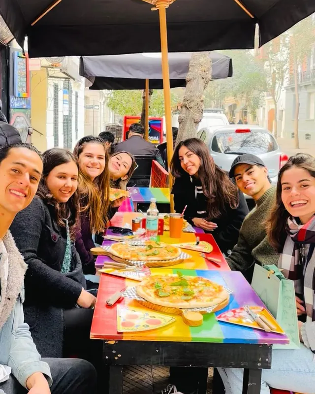 Grupo de personas sonriendo en una mesa arcoíris de la terraza, con dos pizzas